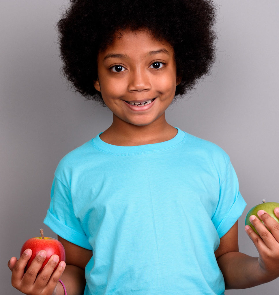 an image of a young african american little girl holding two apples in each hand standing in front of a gray backdrop